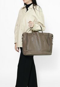 Borsa tote in pelle marrone con superficie testurizzata, due manici e lati con zip, tenuta da una persona in una camicia color crema.
