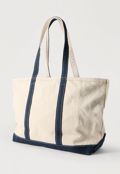 Μπεζ υφασμάτινη τσάντα ώμου με σκούρες μπλε λαβές και περιθώριο, όρθια σε απλό ανοιχτό φόντο.