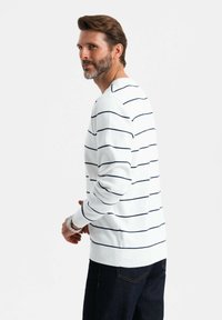 Suéter blanco de punto con rayas horizontales en navy, cuello redondo, puños y dobladillo acanalados, combinado con jeans oscuros. El modelo está ligeramente de lado.