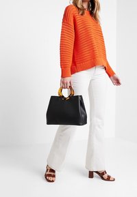 Femme portant un pull en tricot orange, un pantalon blanc évasé et des sandales à talons marron, tenant un sac à main noir avec des poignées en écaille.