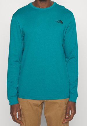 Mężczyzna w morskim, długim rękawie z logo North Face i beżowych spodniach, stojący na jednolitym białym tle.