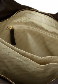 Interno di borsa in pelle marrone con texture liscia. Presenta una tasca con cerniera e due scomparti aperti, foderati con tessuto beige e motivo con il logo del marchio.
