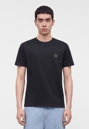 Jeune homme aux cheveux courts et noirs portant un t-shirt noir avec un petit logo rond sur la poitrine gauche et un jean bleu clair.