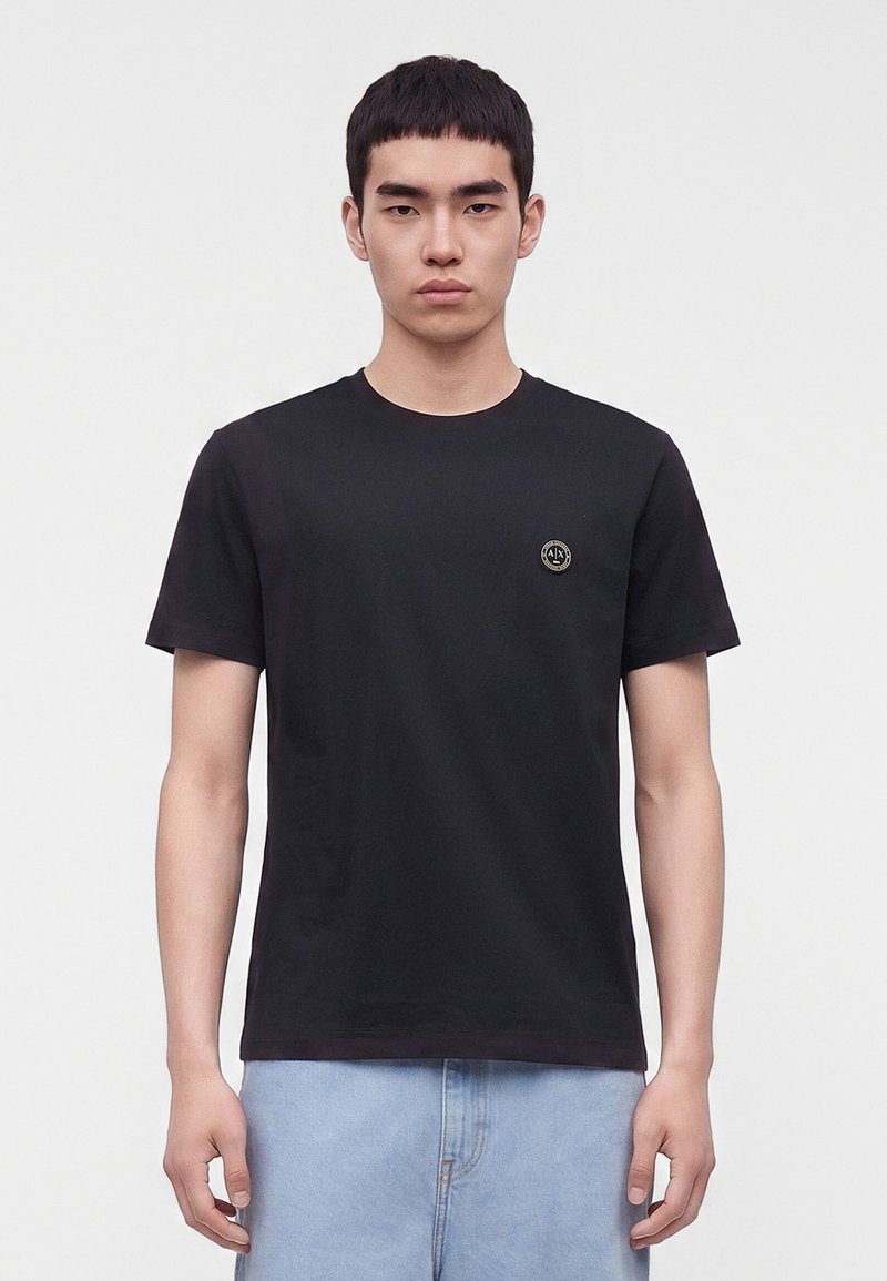 Jeune homme aux cheveux courts et noirs portant un t-shirt noir avec un petit logo rond sur la poitrine gauche et un jean bleu clair.