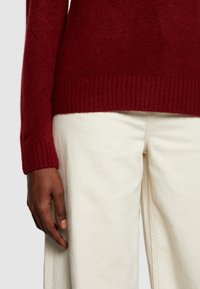 Burgundy neulepaita, jossa on ribattuja hihansuita, yhdistettynä kermanvärisiin housuihin. Kangas vaikuttaa pehmeältä ja teksturoidulta, ja siinä on rento istuvuus.