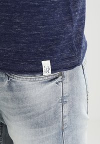 Camisa de manga corta azul marino con un acabado texturizado, combinada con vaqueros desgastados de un azul claro. Etiqueta visible en el dobladillo de la camisa.