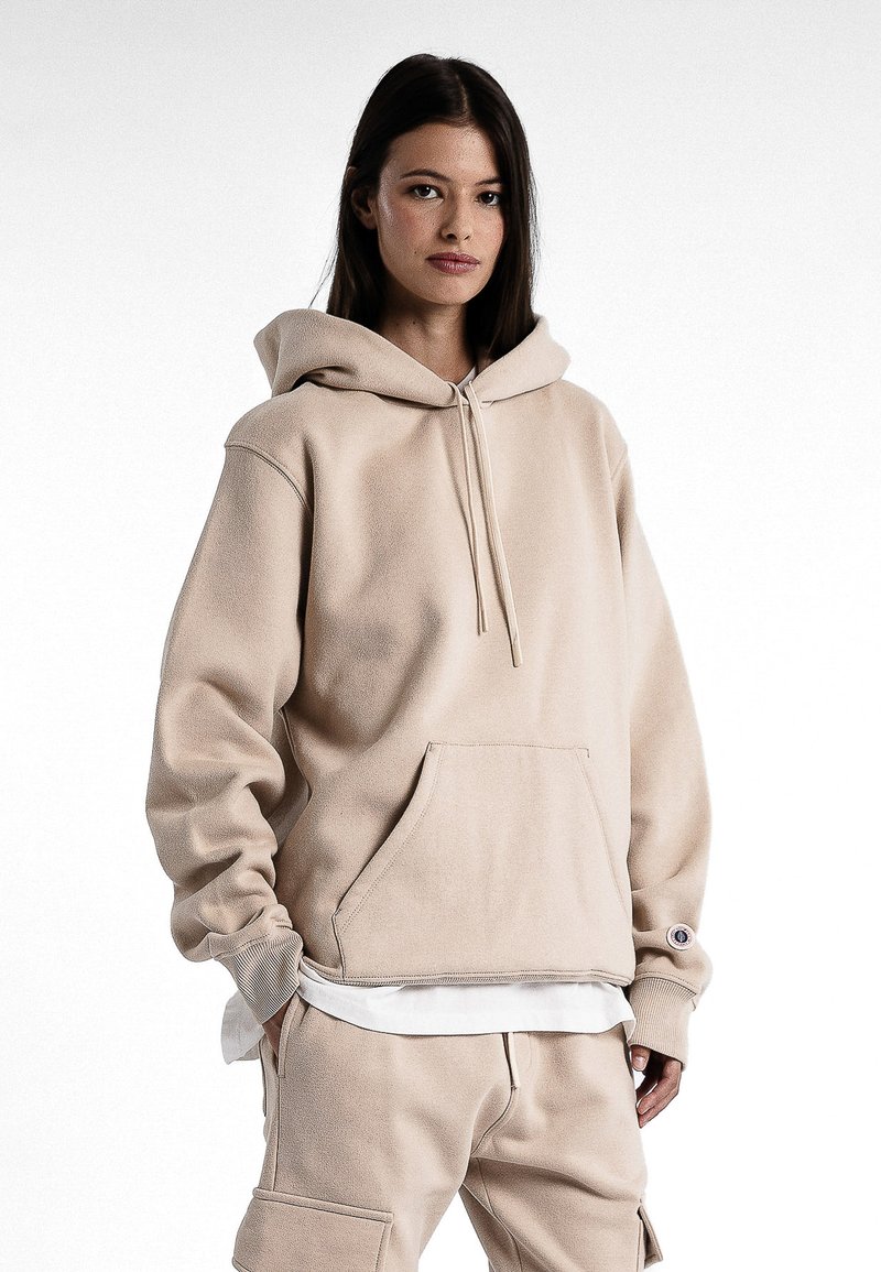 Haut à capuche beige oversize en tissu doux, avec une poche kangourou, un cordon de serrage à la capuche et des poignets côtelés. Porté par-dessus une chemise blanche.