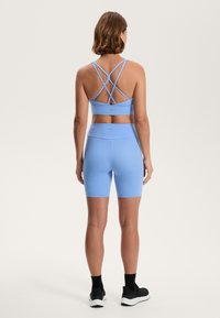 Svetlo modra športna nedrčka s prečnimi naramnicami in ujemajočimi se kolesarskimi hlačami. Gladek, raztegljiv material, oblikovan dizajn in visok pas.
