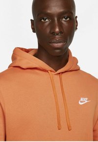 Felpa arancione in morbido tessuto, dotata di cappuccio con coulisse e del logo Nike bianco sul petto. Dettaglio ravvicinato del colletto.