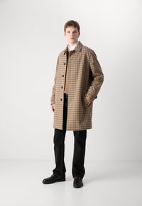 Herrenmantel in Braun und Beige mit Hundszahn-Muster, mit Kragen, Knopfverschluss und Seitentaschen, getragen über einem braunen Pullover und schwarzen Hosen.