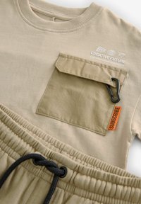 Beige bomull t-shirt med bröstficka, med en svart klämma, och matchande joggers med elastisk midja och en orange etikett som säger "SUBSCRIBE."
