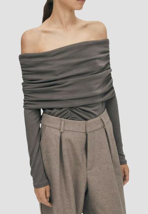 Donna che indossa un top grigio a maniche lunghe arricciato e scollato sulle spalle, con pantaloni beige plissettati a vita alta, su sfondo semplice.