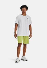 Hvid T-shirt med korte ærmer og sort logo, parret med lysegrønne shorts. Model bærer hvide sportsko med grå detaljer.