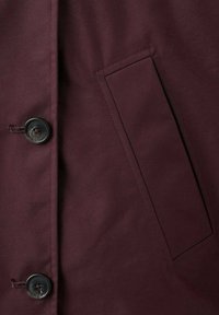 Primer plano de un abrigo de tela burdeos oscuro que muestra dos botones negros y un bolsillo de ojal rectangular en el lado derecho.