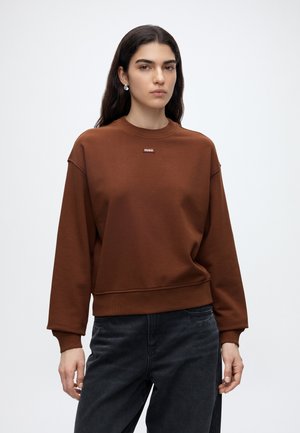 Kvinde med langt mørkt hår iført perleøreringe, en brun sweatshirt med "HUGO" logo og sorte jeans mod en ensfarvet hvid baggrund.