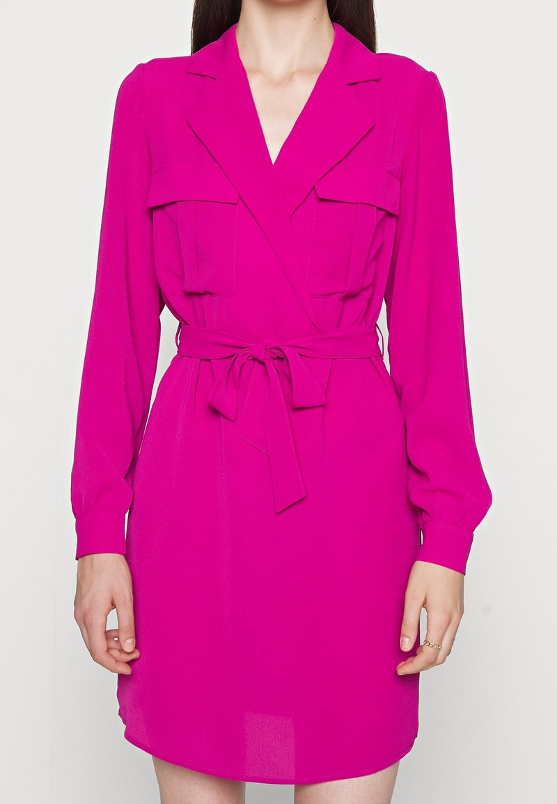 Robe fuchsia à manches longues, avec un col et deux poches avant. Elle possède une ceinture à nouer à la taille et un tissu lisse et léger.