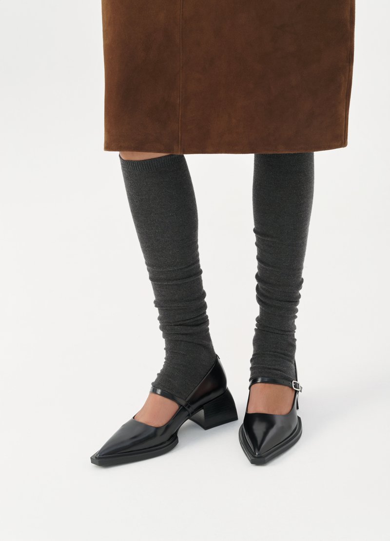 Schwarze Lackleder-Schuhe mit spitzem Zeh, niedrigem Blockabsatz und Knöchellriegel, kombiniert mit dunkelgrauen Rippstrümpfen.