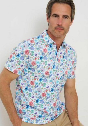 Man draagt een korte mouwen, met knopen versierde bloemenshirt met rode, blauwe en groene bloemen, gecombineerd met beige broek, zittend tegen een effen achtergrond.