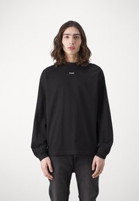 Black long-sleeve sweatshirt featuring a crew neck and the logo "HUGO" in white on the chest. Fabric appears smooth with a relaxed fit.