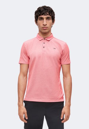Uomo che indossa una polo rosa a maniche corte con bottoni neri e piccolo logo nero su petto e manica, in piedi contro uno sfondo semplice.