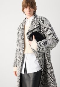 Abrigo de mezcla de lana con un patrón de espiga en blanco y negro, silueta larga, solapas anchas y grandes botones; combinado con un bolso de mano negro.
