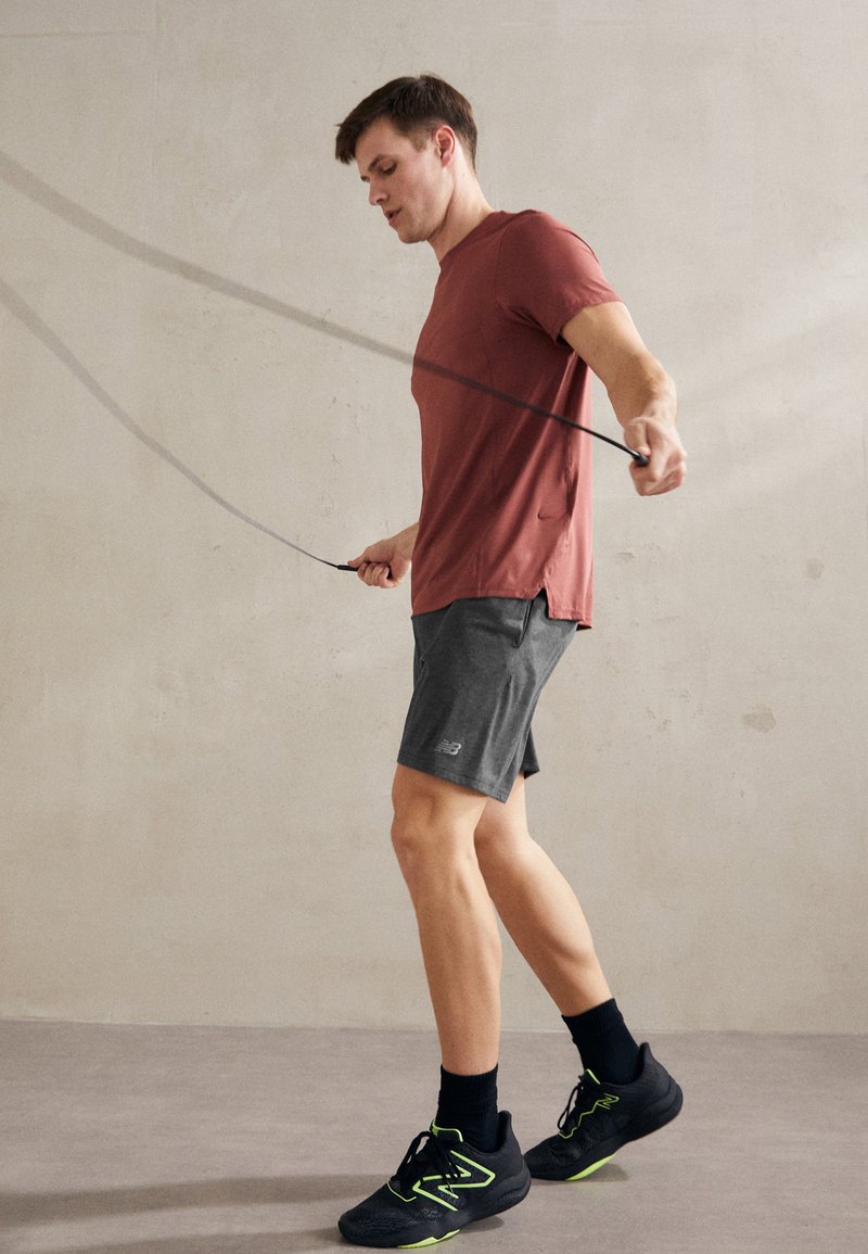 Homme athlétique portant un t-shirt rouille et un short noir en train de sauter à la corde, chaussé de baskets noires avec des accents verts, sur un fond neutre.
