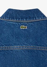 Denim jacket featuring a classic collar, blue color, orange stitching, and a small embroidered crocodile logo on the back.