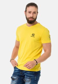 Gelbes Baumwoll-V-Ausschnitt-T-Shirt mit einem schwarzen Logo auf der linken Brust und kurzen Ärmeln. Glatte Textur, taillierter Schnitt mit lässigem Look.
