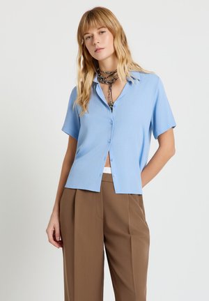 Femme aux cheveux blonds portant une chemise boutonnée bleu clair à manches courtes, un pantalon large marron et des colliers perlés en couches.