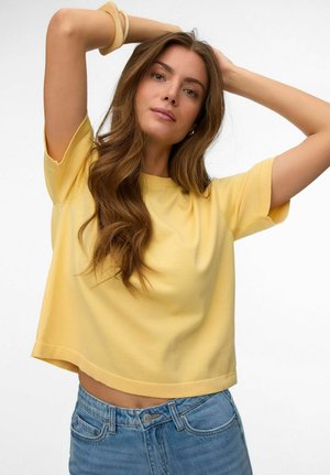 Jeune femme aux longs cheveux bruns portant un haut jaune à manches courtes et un jean bleu, posant avec les mains derrière la tête.