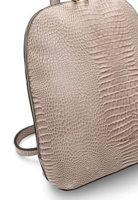 Sac à dos beige avec texture crocodile embossée, fermeture éclair visible et petit logo de marque doré sur la partie inférieure.