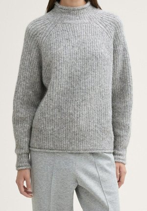 Frau trägt einen lockeren, gerippten grauen Strickpullover mit langen Ärmeln und passende graue Hosen, steht vor einem schlichten Hintergrund.