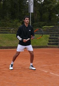 Jogador de ténis masculino com jaqueta preta da adidas, calções brancos e sapatos com detalhes amarelos, segurando uma raquete de ténis num campo de terra batida.