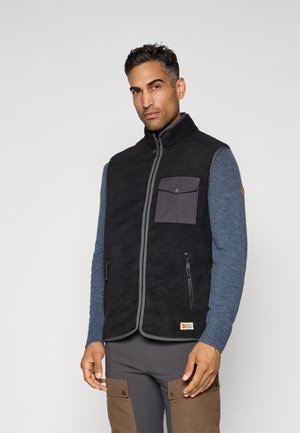 Man in een zwart fleece vest met rits en zak, een blauw gebreid shirt met lange mouwen en grijze broek met bruine cargozakken, staande tegen een witte achtergrond.