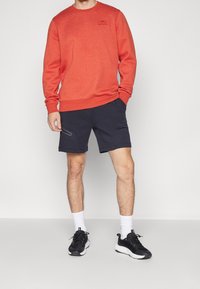 Sudadera roja con cuello redondo y puños acanalados, combinada con pantalones cortos azul marino con bolsillos con cremallera. Se lleva con zapatillas negras y calcetines blancos.