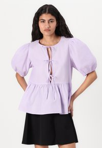 Lavendelbluse med puffærmer, bånd-front detalje og peplum kant. Glat tekstur, letvægtsstof, parret med en sort nederdel.