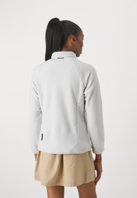 Giacca in fleece grigio chiaro con colletto alto, maniche raglan e orlo dritto. Presenta un piccolo etichetta nera in basso a sinistra.