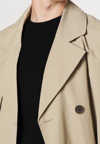 Hellbeiger Trenchcoat mit großem Kragen und einem einzigen dunklen Knopf, getragen über einem schwarzen Strickpullover, der eine glatte Stoffstruktur zeigt.