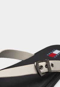 Μαύρο σανδάλι με λευκό ρυθμιζόμενο λουράκι που διαθέτει μαύρη μεταλλική αγκράφα και μικρό κόκκινο, λευκό και μπλε λογότυπο στη σόλα.
