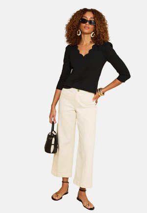 Žena s kučeravými vlasmi nosiaci čierny top so zvlnenými okrajmi, krémové široké nohavice, čierne sandále, slnečné okuliare, zlaté kruhové náušnice, náramky a držiaca čiernu kabelku.