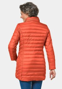 Oranje pufferjack met een hoge kraag en horizontale verstellingen, voorzien van een ritssluiting, tot halverwege de dijen reikt en lange mouwen heeft.