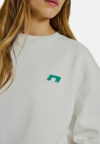 Biely sveter s zeleným logom a dvoma horizontálnymi pruhmi. Dizajn obsahuje rebrovaný okrúhly výstrih a mäkkú, štruktúrovanú tkaninu.