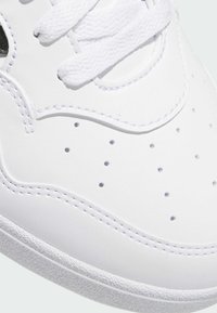 Białe sneakersy wykonane z gładkiego materiału syntetycznego, z perforowanymi detalami, płaskimi sznurowadłami oraz teksturowaną gumową podeszwą.