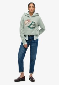 Donna in piedi che indossa una felpa con cappuccio verde chiaro con stampa grafica, una maglietta a righe sotto, jeans blu con risvolto e mocassini scuri.