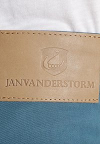 Textiletikett mit hellbraunem Kunstleder, geprägt mit einem Schild- und Bootdesign, auf blauen Stoff mit diagonaler Textur aufgenäht.