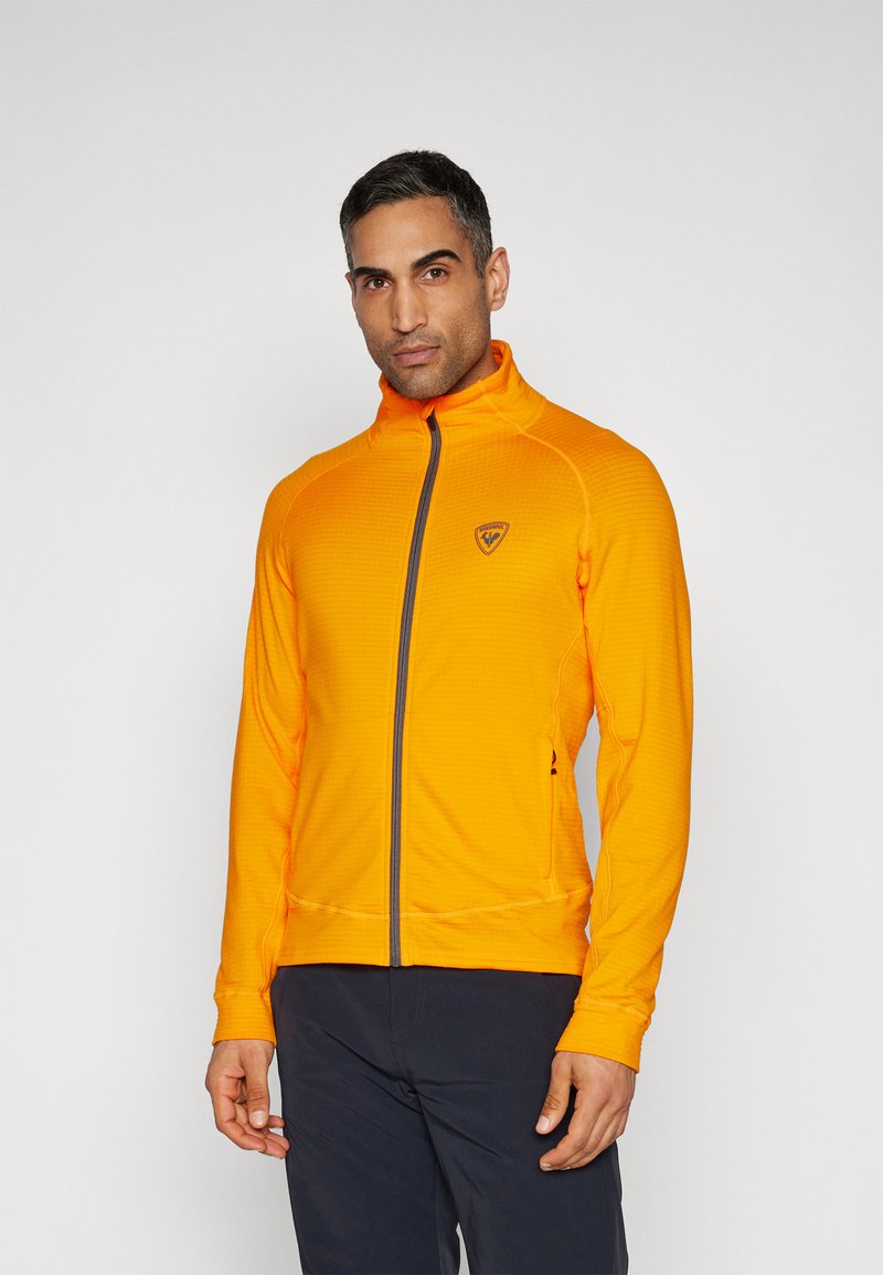 Oranje ritsjack met een gestructureerd patroon, hoge kraag en grijze accenten. Bevat een logo-opdruk op het linker borstgebied.