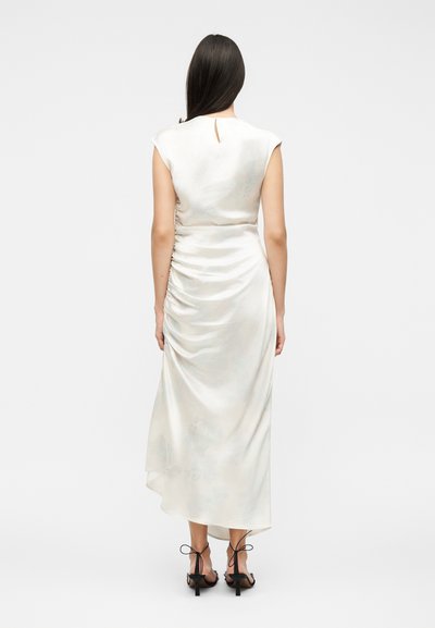 Γυναίκα με μακριά σκουρόχρωμα μαλλιά, που φοράει ένα αμάνικο, ριγέ, μίντι φόρεμα από σατέν εκρού και μαύρα ψηλοτάκουνα με λουράκια, γυρισμένη με την πλάτη σε ένα απλό φόντο.