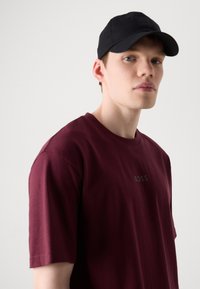 Bordeauxfarbendes Baumwoll-T-Shirt mit Rundhalsausschnitt und kurzen Ärmeln, versehen mit einem schwarzen "BOSS"-Logo auf der Brust. Getragen mit einer schwarzen Kappe.