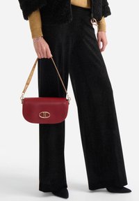 Borsa a mano in pelle rossa con chiusura in metallo dorato, abbinata a pantaloni neri larghi con sottili scintillii e a una giacca nera con texture.