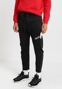 Persoon in zwarte joggers metkoord en witte tekst op de dij, witte sokken, zwart-witte sneakers en een rode hoodie.